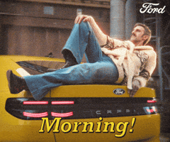 Sponsored gif. Man poses dramatically along the back of a yellow vehicle. He tilts his head forward and gives an intense look with one eyebrow arched. Text reads, “Morning!”