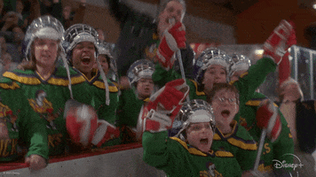 Movie gif. The Mighty Ducks hockey team from The Mighty Ducks erupts into loud and excited celebration alongside the edge of a rink.