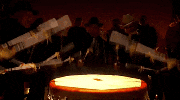 Oscars 2024 gif. Performing 'Wahzhazhe (A Song For My People)' from Killers of the Flower Moon, men adorning all black and wearing cowboy hats stand around a tribal drum and beat on it in unison. They're shrouded in the shadows while an ethereal glow hits the drum. 
