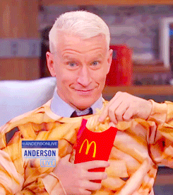 Celebrity gif. Anderson Cooper is wearing a French fry shirt and he has a crazed expression on as he shoves a handful of McDonald's fries into his mouth.