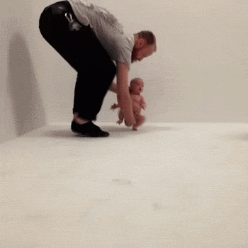 Video gif. A dad holds a diaper-wearing baby by its neck and bottom and hustles it across the floor. Its legs move quickly trying to keep up.