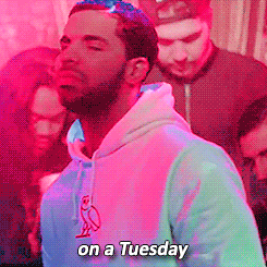 Music video gif. Young Drake is rapping in a club and there are people sitting around him, grooving and drinking. He says, "On a Tuesday," while looking at us and pointing. 