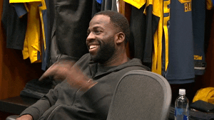 Sports gif. Draymond Green from the Golden State Warriors is sitting in the locker room listening to others talk. He laughs and looks delighted.