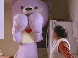 Video gif. A person dressed in a purple teddy bear costume with a giant head fumbles something in his arms, then holds his arms up in front of his face in embarrassment. Text, “OOPS.”