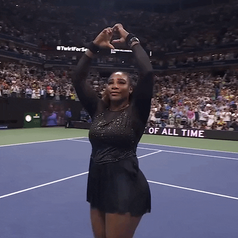 Sports gif. Serena Williams wears a black sparkling outfit and holds her hands over her head in a heart shape as she walks on a tennis court.