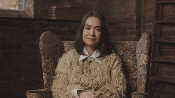 Video gif. Woman sitting in a chair raises her eyebrows and shrugs her shoulders, throwing her hands up as if to say, “I don’t know.”