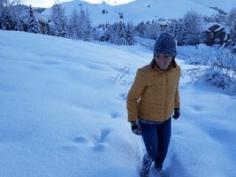 Video gif. A woman jumps backwards with her hands in the air into a pile of snow and lands deep in the pile.