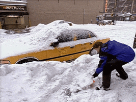 TV gif. Conan O'Brien is outside on a city street in the snow wearing winter gear. He laughs as he uses a shovel to pick up a pile of snow and then chucks it onto a taxi cab already snow covered windshield.