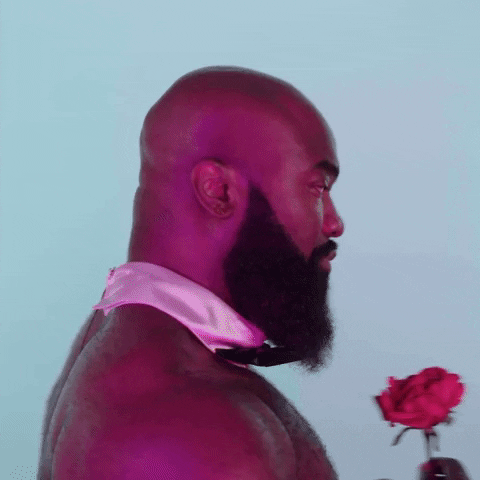 Video gif. Man with no shirt on, but a collar and bow tie, turns around with a rose in his hand. He first looks serious, but then his face melts into a sweet smile as he says, “I love you.”