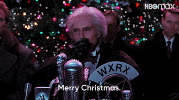 Movie gif. Christopher Walken as Max in Batman Returns stands at a microphone making a speech with a large outdoor Christmas tree behind him, as he waves and says, "Merry Christmas."