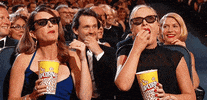 Celebrity gif. Amy Poehler and Tina Fey at the Emmys sitting in the audience with 3D glasses on and a tub of popcorn. They munch and stare at the stage, engrossed in the live action scene they're watching.