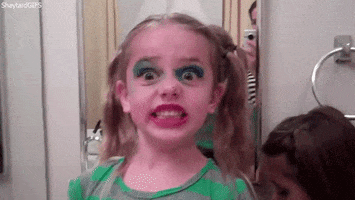 Video gif. A toddler with pigtails has done her own makeup and her face is painted with big blue eyeshadow and red lips. She looks pleased and she gives us a big grin.
