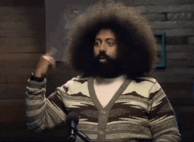 Celebrity gif. Reggie Watts does the cuckoo sign, twirling his finger at his ear and making an O with his mouth as he raises his eyebrows.  