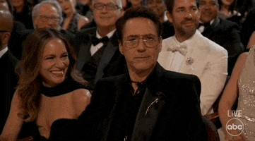 Oscars 2024 GIF. Robert Downey Junior, seated at the Oscars, looks at us and makes a scene of shrugging his shoulders up to his ears in uncertainty, then dropping them and batting his eyelashes in postured innocence.