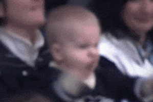 Video gif. A baby raises both fists in the air as he yells in immense excitement. 