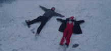 Music video gif. An aerial shot of Ed Sheeran and Elton John laying in a snow field, making snow angels.