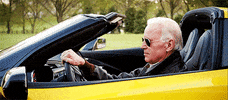Politics gif. Joe Biden in sunglasses turns to us and grins from behind the wheel of a banana-yellow Corvette in a parody video about life in the White House.