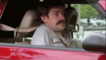 The Office gif. John Krasinski as Jim in The Office sits in a car and wears a fake mustache, looking suspiciously out the window. He lowers the car seat until he is out of sight.