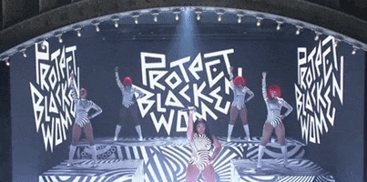 SNL gif. Megan Thee Stallion is on stage at SNL, raising her microphone in the air as she stands in front of a backdrop that says, "Protect Black Women." Four background dancers stand on platforms and do the same pose, all wearing bright red wigs and black and white spiral leotards. 