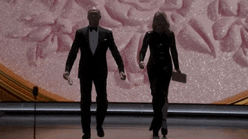 Oscars 2024 GIF. Michael Keaton and Catherine O'Hara walk on stage but Keaton walks way too fast and O'Hara stops and calls out after him in faux frustration. Keaton immediately turns around and grins sheepishly while he waits for her to catch up. He holds her hand when she approaches and they finish the walk to the podium together.