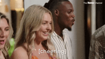 Reality TV gif. Contestant on "Married at First Sight" smiles and claps her hands together, doing a little shoulder dance as other contestants cheer behind her. Subtitle audio indicates, "Cheering."