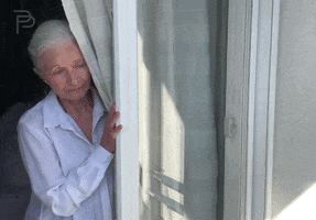 Video gif. An elderly woman looks out a window from behind a curtain, seeming lonely and sad. Sparkling pink text moves down the screen, "I miss you."