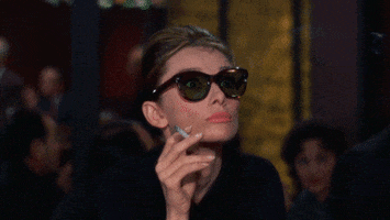 Movie gif. Aubrey Hepburn as Holly Golightly in Breakfast at Tiffany’s wears sunglasses and holds a smoking cigarette in between her fingers. Her eyes widen behind her tinted lens and her eyebrow raised in shock. She slides her sunglasses down her nose and looks over her glasses up at someone.