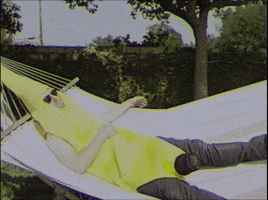 Video gif. Man in a banana costume is laying in a hammock. He has sunglasses on and he lazily lifts a hand up while the text, "Bored?" flashes on the screen.