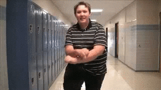 Video gif. A student in high school is standing in the hallway and he attempts to dance Psy's Gangnam Style dance. He looks jolly as he jumps up and down.