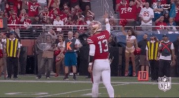 Sports gif. Brock Purdy of the San Francisco 49ers walks in slow motion across the field with one hand up making the sign for number one as fans and cheerleaders jump up and celebrate. 