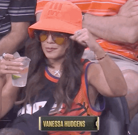 Celebrity gif. Vanessa Hudgens is at the 2021 NBA Finals and she wears a bucket hat and a jersey while holding a drink. She puts her hand in the air and does a stank face as she starts dancing along to the music.