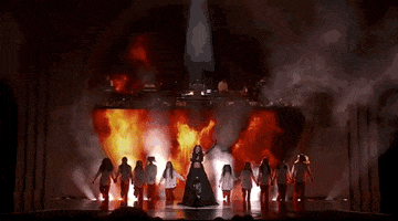 Oscars 2024 GIF. Wide shot of Becky G and a girls' choir performing The Fire Inside from Flamin' Hot. Becky G and the girls walk powerfully forward on the stage and the girls are all wearing a white top and red pants. Becky G pumps her fist in the air as flames shoot up in the background.