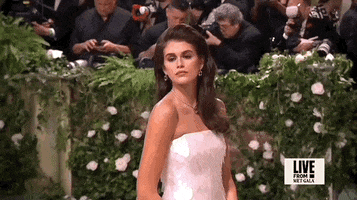 Met Gala 2024 gif. Close up of Kaia Gerber wearing a white strapless floor-length Prada featuring a paillette pattern of large white sequins with a straight neckline. Her hair is styled in a side-part bouffant. This is paired with a delicate diamond pendant necklace.