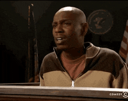 TV gif. Comedian Dave Chappelle sits in a courtroom stand. He moves his head, lifting one eyebrow up in confusion. He says, “huh?” 