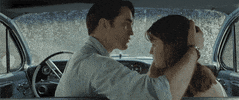 Movie gif. We watch from the backseat of a car in the rain while Robert Pattinson as Preston in The Devil All the Time gently pulls Mia Wasikowska as Helen's head toward him and kisses her forehead.