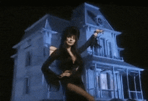 Movie gif. Cassandra Peterson as Elvira in Elvira: Mistress of the Dark. She stands with a hand on her hip and a hand in the air, displaying a two story haunted house while lightning flashes in the back.