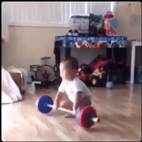Video gif. A small baby stands straight up, holding a foam weightlifting barbell toy over his head with a big smile on his face. He slams it down on the ground and then looks down to the ground and screams like he’s full of adrenaline. 