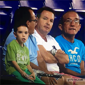 Sports gif. A little boy and three men sit in the audience at a stadium. They suddenly notice they're on the jumbotron and start waving their arms. The boy screams and throws an arm up, then starts dancing with his tongue out, and pulls up his shirt as he moves his hips. 
