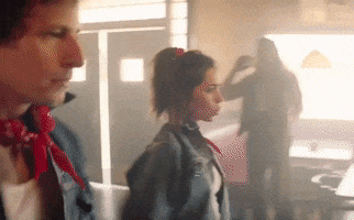 Movie gif. Andy Samberg as Nyles and Cristin Milioti as Sarah in "Palm Springs" do a coordinated hip-swinging dance in a dusky daylit bar, wearing matching jean jackets, white T's, black jeans, and red scarves.