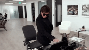 Reality TV gif. Kris Jenner on Keeping Up With The Kardashians stands over a desk and closes up a laptop. She has blacked out sunglasses on and she looks suspicious as she goes to grab something on the desk. 