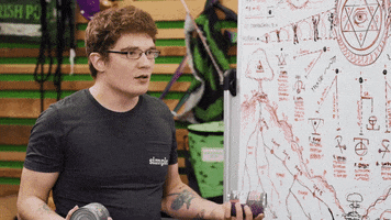 Video gif. A man is standing in front of a whiteboard filled with notes and he looks very upset as he asks, "What is happening here?"
