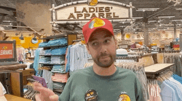 Video gif. A man stands in the middle of a Bass Pro Shop. He shakes his head and holds two tightly crossed fingers up. He says, “Fingers crossed,” and walks away. 