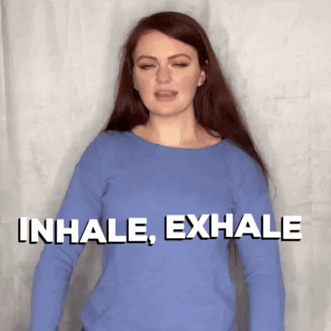 Gif of a white woman with long dark hair wearing a blue top raising her hands up and down in front of her saying 'inhale, exhale'