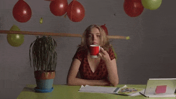 Video gif. A woman looking unamused sips from a coffee cup near a dying desk plant as she gazes ahead with a sigh. A hand holds out a long pole and pops balloons that hang behind her as papers drop from above her.
