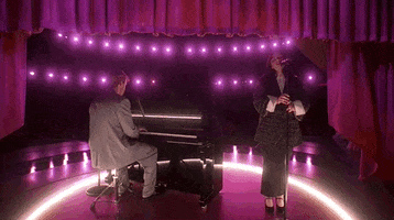 Oscars 2024 GIF. Billie Eilish and Finneas performing "What Was I Made For" on stage at the Oscars. Finneas is on the piano and Eilish stands next to him, softly holding the microphone. Finneas wears a grey suit and Eilish wears a long skirt with an oversized Chanel blazer, in the classic Chanel tweed. She has an exaggerated white collar and sleeves, which fold over the jacket's arms. The background is bathed in Barbie pink. 