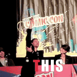 Celebrity gif. Tom Hiddleston sits on a panel at New York Comic Con. He holds his arm up, stretching high up while pointing his finger. He twists his body around. Text, “This.”