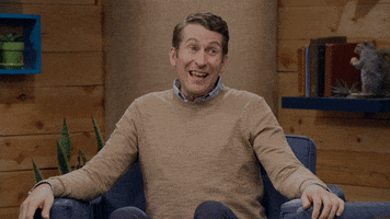 Video gif. Scott Aukerman from Comedy Bang Bang is recording a podcast video. He sits on a couch and raises his eyebrows with an open mouth, gesturing around but looking confused and unsure how to answer.