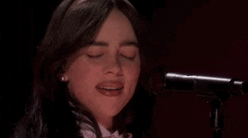 Oscars 2024 GIF. Close up shot of Billie Eilish performing "What Was I Made For" on stage at the Oscars. Eilish sings the last word of the song and pulls away from the microphone, looking at Finneas and grinning. 
