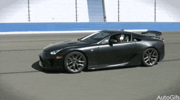 Video gif. Black Lexus LFA car speeds across a racetrack, the sun glinting off its windows.