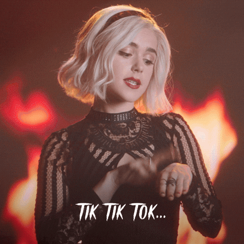 Music video gif. Kiernan Shipka as Sabrina the Chilling Adventures of Sabrina's music video Straight to Hell, taps her wrist as she stands in front of fire. She looks at us as she sings, "Tik tik tok..."